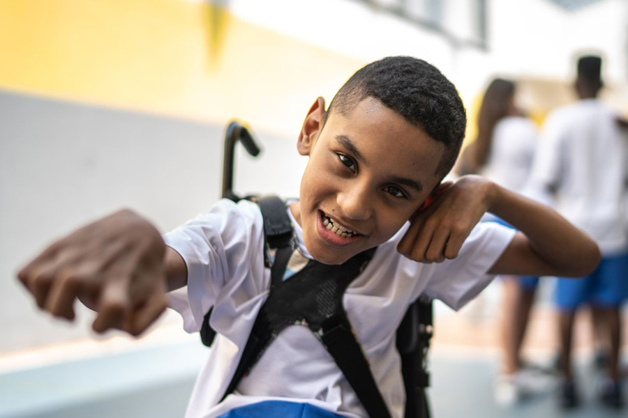 Portrait of a Brazilian student in a sports court. He is a wheelchair user, wearing a white shirt and blue shorts. In the background, other students are wearing a similar sports uniform.