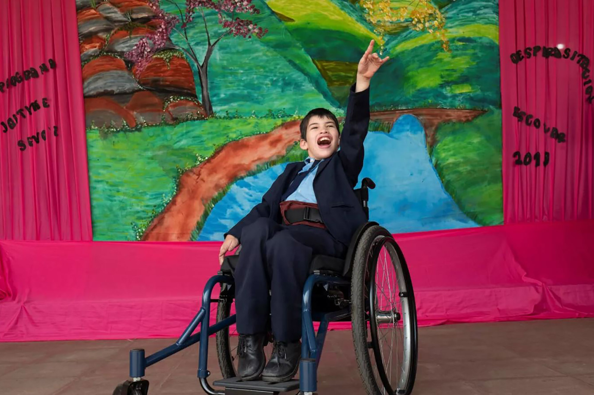 A young boy in a wheelchair is performing on a stage and pointing up.