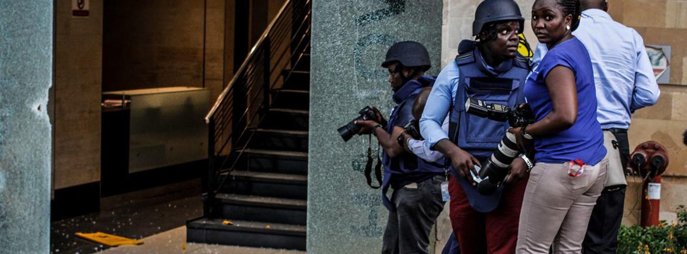 Journalists in protective gear in front of building with broken glass entrance.