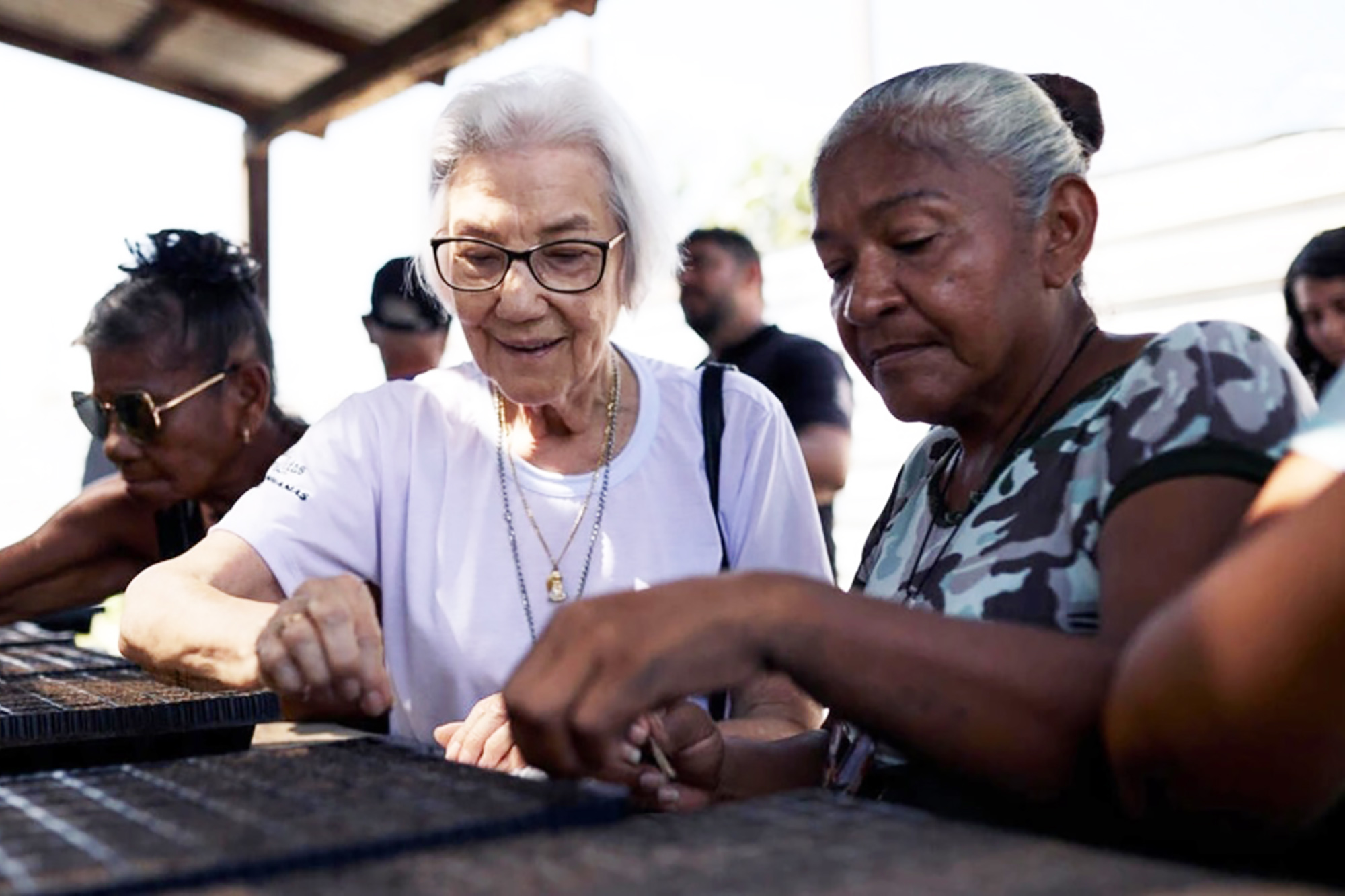 Two women are sowing seeds in small pots placed on a table.