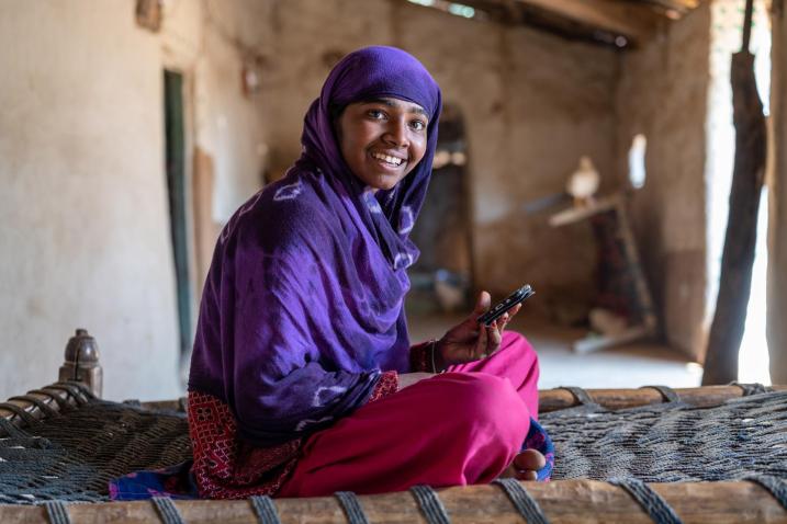 A girl wearing a purple head scarf is sitting on a bed in an indoor setting. 