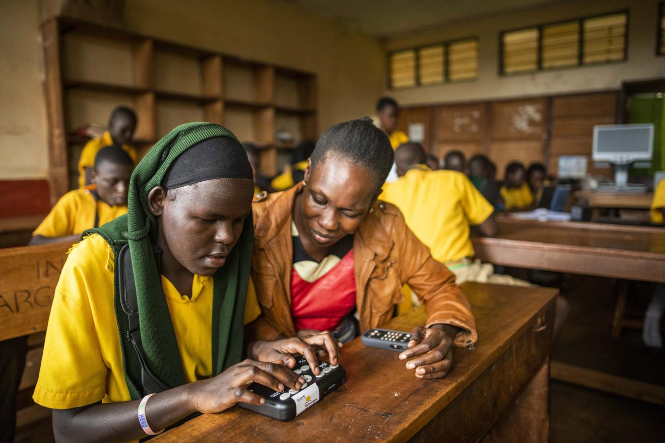 A teacher helping a student to use an optic braille reader in a classroom.