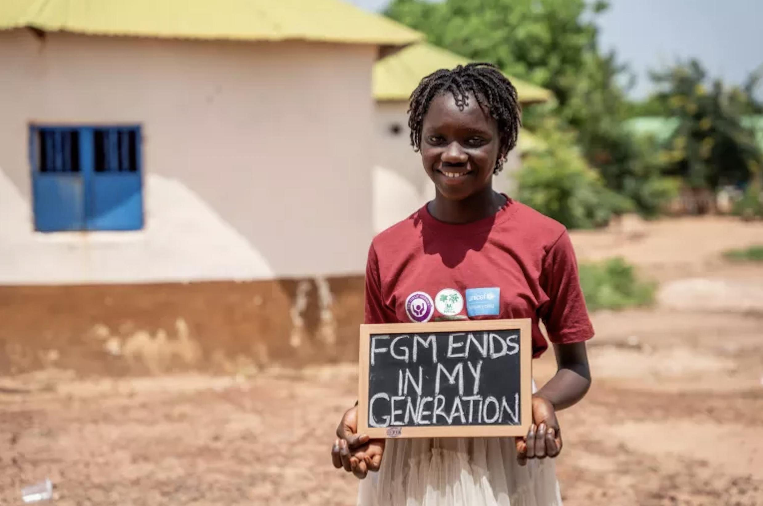 A photograph showing a young woman smiling at the camera and holding a chalkboard sign that reads 'FGM ENDS INN MY GENERATION'